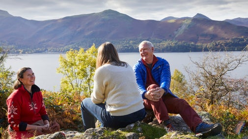 A trio of people sit looking out on a view over a lake from a mountainside in the Lake District.